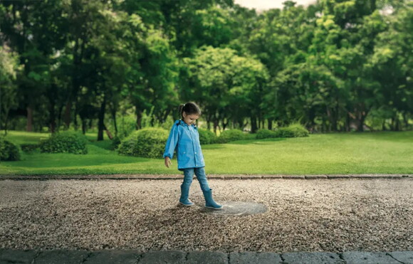 Niña con impermeable azul y botas de lluvia, mirando un charco en un camino de grava en un parque verde y frondoso Niña con impermeable azul y botas de lluvia, mirando un charco en un camino de grava en un parque verde y frondoso
